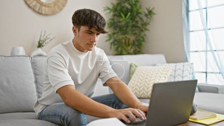 Téléchargez les photos : Jeune étudiant hispanique concentré, absorbé par l'éducation en ligne, assis sur un canapé confortable dans le salon, concentré sur l'écran d'ordinateur portable, relaxant à la maison, entouré de technologie - en image libre de droit