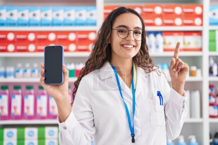 Photo for Young hispanic woman working at pharmacy drugstore showing smartphone screen smiling and laughing hard out loud because funny crazy joke. - Royalty Free Image