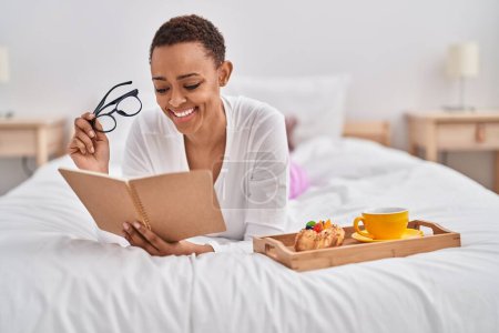 Téléchargez les photos : Femme afro-américaine prenant le petit déjeuner livre de lecture dans la chambre - en image libre de droit