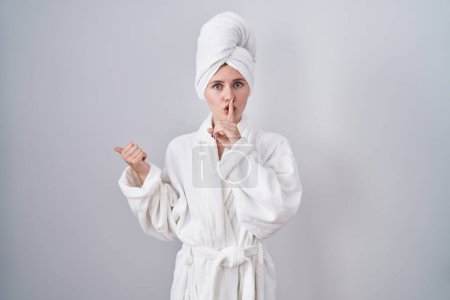 Photo for Blonde caucasian woman wearing bathrobe asking to be quiet with finger on lips pointing with hand to the side. silence and secret concept. - Royalty Free Image