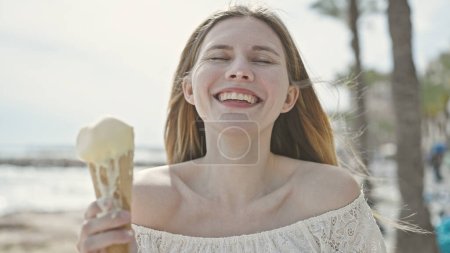 Foto de Joven mujer rubia turista sonriendo confiado sosteniendo helado en la playa - Imagen libre de derechos