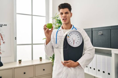 Photo for Young hispanic doctor man holding scale at dietitian clinic clueless and confused expression. doubt concept. - Royalty Free Image