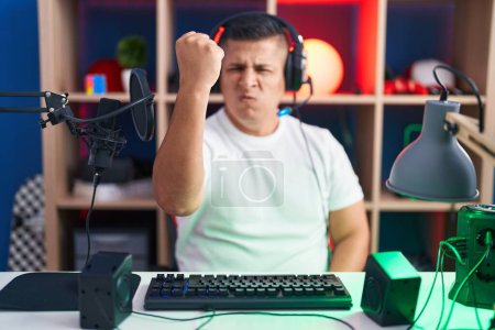 Foto de Joven hombre hispano jugando videojuegos enojado y loco levantando el puño frustrado y furioso mientras gritaba con ira. rabia y concepto agresivo. - Imagen libre de derechos