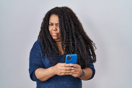 Foto de Plus size hispanic woman using smartphone typing message skeptic and nervous, frowning upset because of problem. negative person. - Imagen libre de derechos
