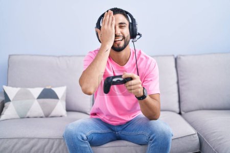 Téléchargez les photos : Jeune homme hispanique jouant contrôleur de tenue de jeu vidéo assis sur le canapé couvrant un oeil avec la main, sourire confiant sur le visage et l'émotion surprise. - en image libre de droit