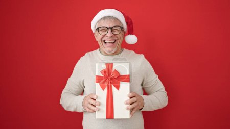 Photo for Middle age grey-haired man holding gift wearing christmas hat over isolated red background - Royalty Free Image