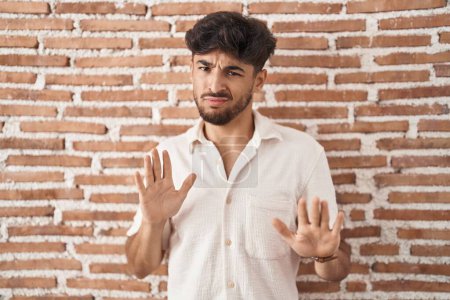 Photo for Arab man with beard standing over bricks wall background moving away hands palms showing refusal and denial with afraid and disgusting expression. stop and forbidden. - Royalty Free Image