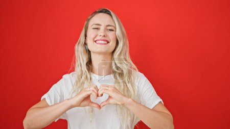 Téléchargez les photos : Jeune femme blonde souriante confiante faisant geste de coeur avec les mains sur fond rouge isolé - en image libre de droit