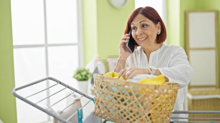 Foto de Mujer de mediana edad hablando en el teléfono inteligente cesta con ropa en la lavandería - Imagen libre de derechos