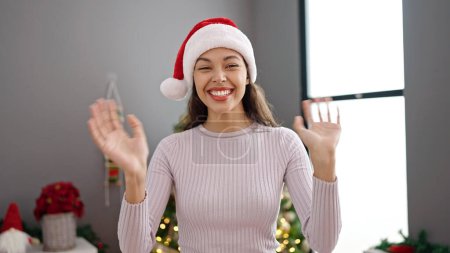 Photo for Young beautiful hispanic woman waving to the camera by christmas tree at home - Royalty Free Image