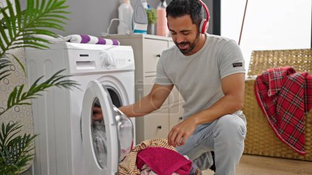 Foto de Joven hispano escuchando música lavando ropa en la lavandería - Imagen libre de derechos