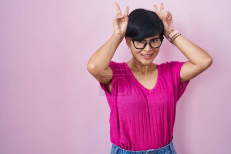 Foto de Joven mujer asiática con el pelo corto de pie sobre fondo rosa posando divertido y loco con los dedos en la cabeza como orejas de conejo, sonriente alegre - Imagen libre de derechos