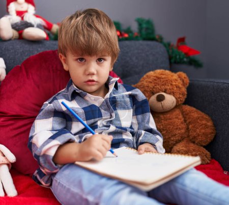 Foto de Adorable niño hispano escribiendo en el cuaderno sentado en el sofá por la decoración de Navidad en casa - Imagen libre de derechos