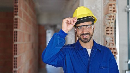Photo for Young hispanic man worker wearing hardhat smiling at construction site - Royalty Free Image