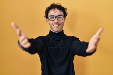 Foto de Hispanic man standing over yellow background looking at the camera smiling with open arms for hug. cheerful expression embracing happiness. - Imagen libre de derechos