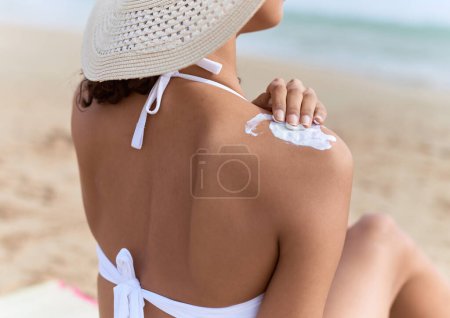 Téléchargez les photos : Jeune femme afro-américaine portant un chapeau d'été et un bikini appliquant de la crème solaire sur l'épaule à la plage - en image libre de droit