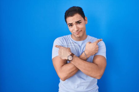 Téléchargez les photos : Jeune homme hispanique debout sur fond bleu pointant des deux côtés avec les doigts, direction différente en désaccord - en image libre de droit