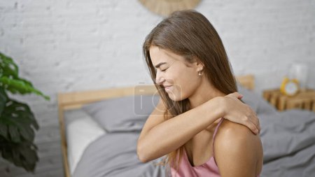 Foto de Mujer hispana joven estresada, hermosa pero preocupada, sentada en la cama en su acogedor dormitorio, sufriendo de dolor de espalda severo, visiblemente incómoda en su pijama ajustado - Imagen libre de derechos