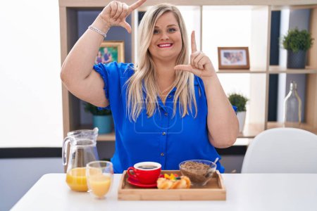 Téléchargez les photos : Femme blanche plus la taille de manger le petit déjeuner à la maison souriant faisant cadre avec les mains et les doigts avec le visage heureux. concept de créativité et photographie. - en image libre de droit