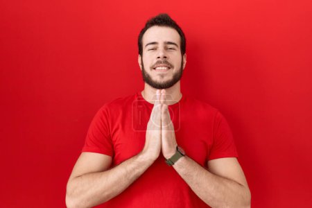 Téléchargez les photos : Jeune homme hispanique portant t-shirt rouge occasionnel priant avec les mains ensemble demandant pardon souriant confiant. - en image libre de droit