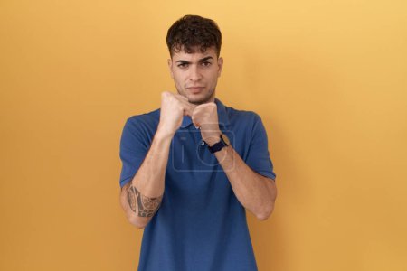 Foto de Young hispanic man standing over yellow background ready to fight with fist defense gesture, angry and upset face, afraid of problem - Imagen libre de derechos