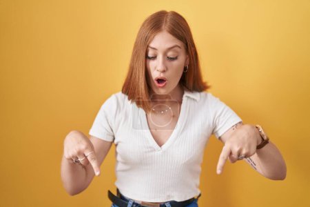 Photo for Young redhead woman standing over yellow background pointing down with fingers showing advertisement, surprised face and open mouth - Royalty Free Image
