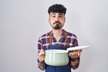 Téléchargez les photos : Jeune homme hispanique avec la barbe portant tablier tenant casserole de cuisson joues gonflées avec drôle de visage. bouche gonflée d'air, attrapant l'air. - en image libre de droit