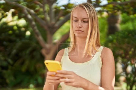 Téléchargez les photos : Jeune femme blonde utilisant un smartphone avec une expression détendue au parc - en image libre de droit