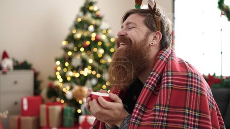 Photo for Young redhead man drinking coffee sitting on sofa by christmas tree at home - Royalty Free Image
