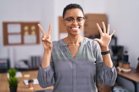 Téléchargez les photos : Femme afro-américaine travaillant au bureau portant des lunettes montrant et pointant vers le haut avec les doigts numéro sept tout en souriant confiant et heureux. - en image libre de droit