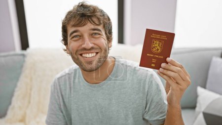 Photo for Smiling young man, confidently holding finland passport, chilling at home on the sofa, ready to embrace his upcoming travel adventure! - Royalty Free Image