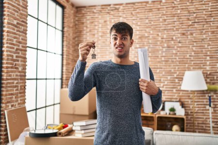 Photo for Young hispanic man holding keys of new home and blueprints clueless and confused expression. doubt concept. - Royalty Free Image