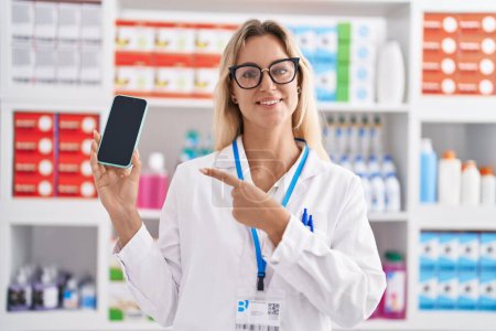 Foto de Young blonde woman working at pharmacy drugstore showing smartphone screen smiling happy pointing with hand and finger - Imagen libre de derechos