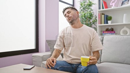Photo for Attractive young hispanic man stretching his neck, savoring morning espresso, enjoying the comfort of home - Royalty Free Image