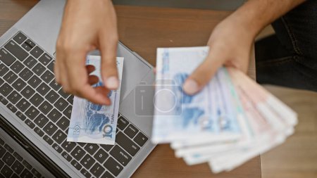 Téléchargez les photos : Jeune adulte assis sur son canapé salon comptant les billets de banque yuan, gérant sa richesse via un ordinateur portable à sa maison confortable - en image libre de droit