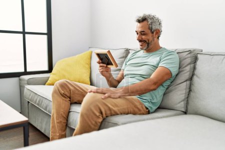 Foto de Hombre de pelo gris de mediana edad que mira la imagen sentado en el sofá en casa - Imagen libre de derechos