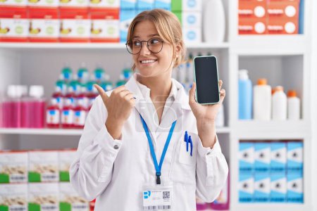 Foto de Joven mujer caucásica que trabaja en la farmacia mostrando la pantalla del teléfono inteligente apuntando el pulgar hacia el lado sonriendo feliz con la boca abierta - Imagen libre de derechos
