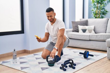 Photo for African american man using smartphone holding kettlebell at home - Royalty Free Image