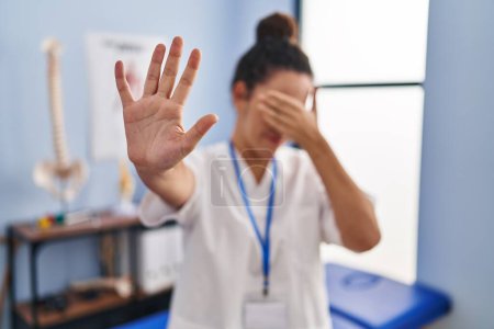 Photo for Young hispanic woman working at rehabilitation clinic covering eyes with hands and doing stop gesture with sad and fear expression. embarrassed and negative concept. - Royalty Free Image