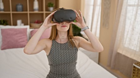 Téléchargez les photos : Jeune femme blonde jouer à un jeu vidéo en utilisant des lunettes de réalité virtuelle assis sur le lit dans la chambre - en image libre de droit