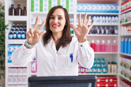 Photo for Middle age brunette woman working at pharmacy drugstore showing and pointing up with fingers number eight while smiling confident and happy. - Royalty Free Image
