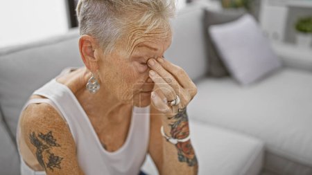Photo for Stressed senior grey-haired woman sitting alone on sofa at home grappling with unseen emotional conflict. - Royalty Free Image