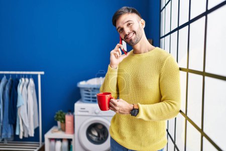 Photo for Young hispanic man talking on smartphone drinking coffee waiting for washing machine at laundry room - Royalty Free Image