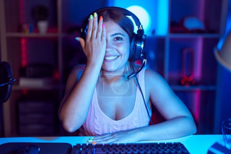 Téléchargez les photos : Jeune femme blonde jouant à des jeux vidéo portant un casque couvrant un oeil avec la main, sourire confiant sur le visage et l'émotion surprise. - en image libre de droit