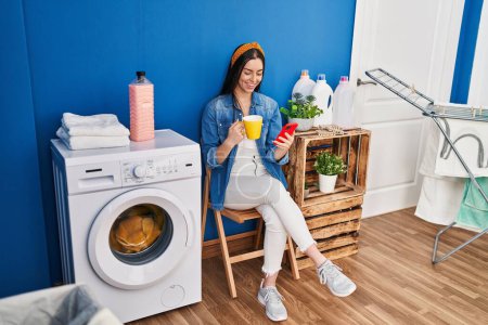 Foto de Joven hermosa mujer hispana usando teléfono inteligente bebiendo café esperando la lavadora en la sala de lavandería - Imagen libre de derechos