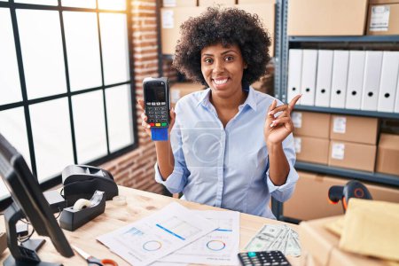 Photo for Black woman with curly hair working at small business ecommerce holding credit card and dataphone smiling happy pointing with hand and finger to the side - Royalty Free Image