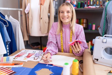 Téléchargez les photos : Jeune femme blonde tailleur écriture sur un bloc-notes tenant chiffon à la boutique de tailleur - en image libre de droit
