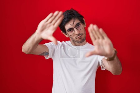 Photo for Young hispanic man standing over red background doing frame using hands palms and fingers, camera perspective - Royalty Free Image