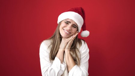 Foto de Joven hermosa mujer hispana sonriendo confiada usando sombrero de Navidad con las manos en la cara sobre fondo rojo aislado - Imagen libre de derechos