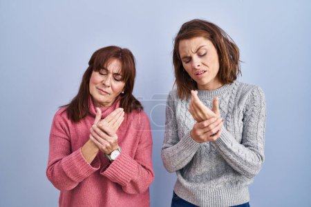 Foto de Madre e hija de pie sobre fondo azul sufriendo dolor en manos y dedos, inflamación de la artritis - Imagen libre de derechos